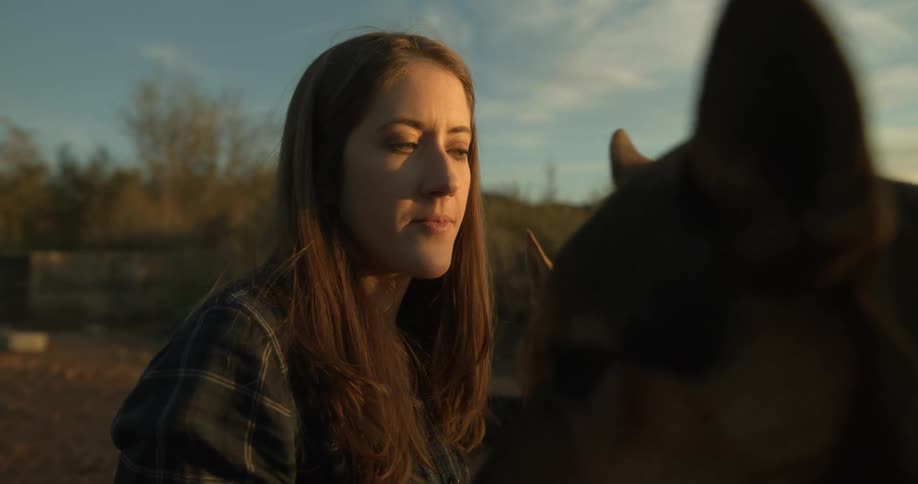 Woman feeds treat to pet dogs crouching in field with hills on horizon