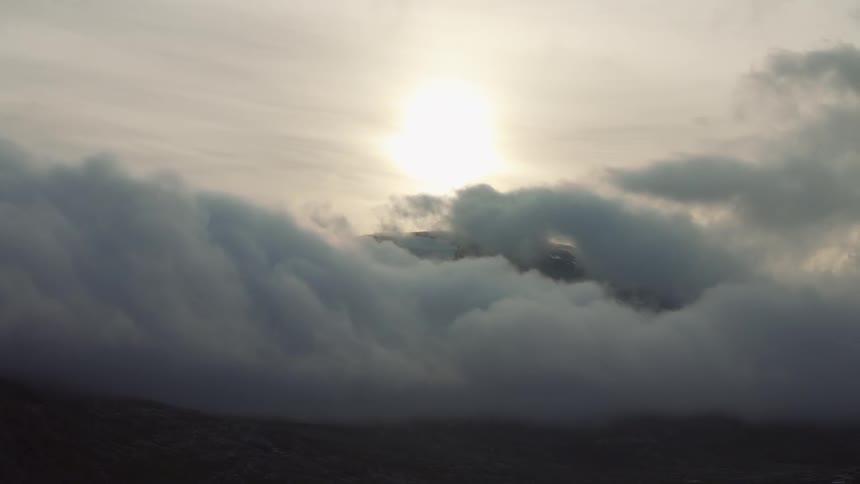 The sun looms over a mountain peak shrouded by clouds