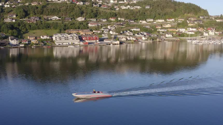 Aerial view of a boat driving over a calm lake near a small coastal town in the mountains