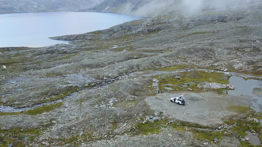An aerial view of a person at the back of a car parked close to a river on flat land