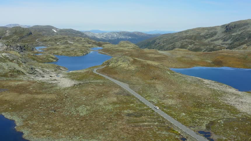 White vehicle drives down highway through rough terrain dotted with lakes