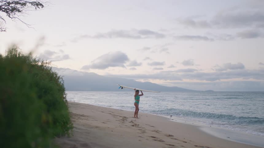 A woman is holding a surfboard on the beach at sunset