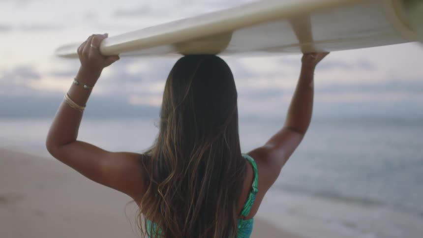 Woman wearing swimsuit carries surfboard on her head as she walks along beach