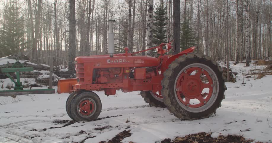 Red tractor sits on snow in yard with leafless trees in the background