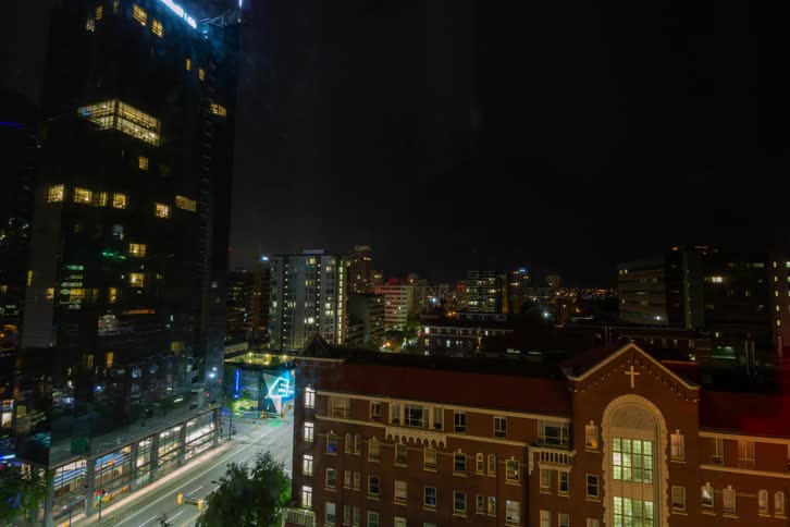 Cars travel on a city street at night in a blur of lights as window lights flicker on and off in the buildings