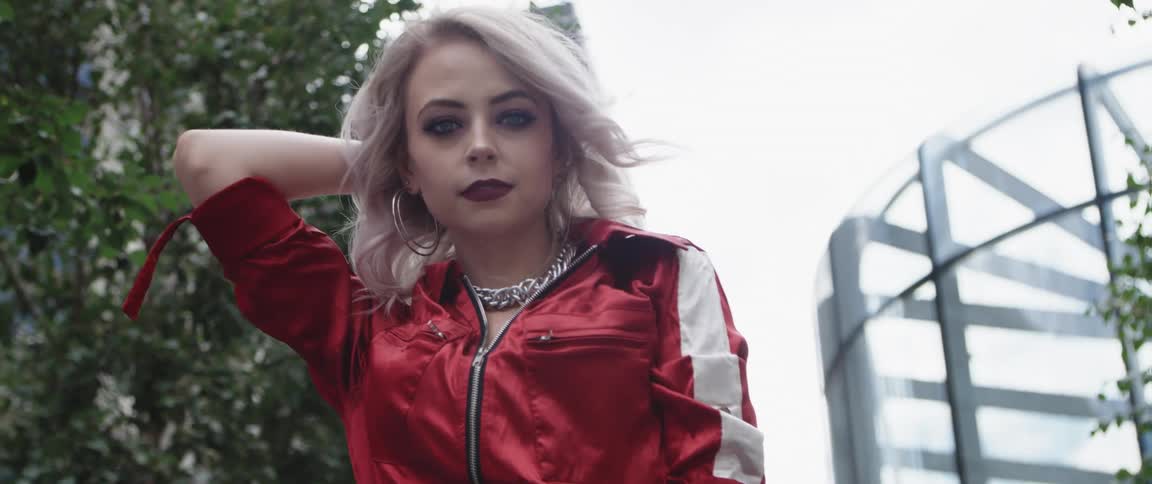 Woman with makeup gray hair hoop earrings necklace and red jacket looks down at camera outdoors