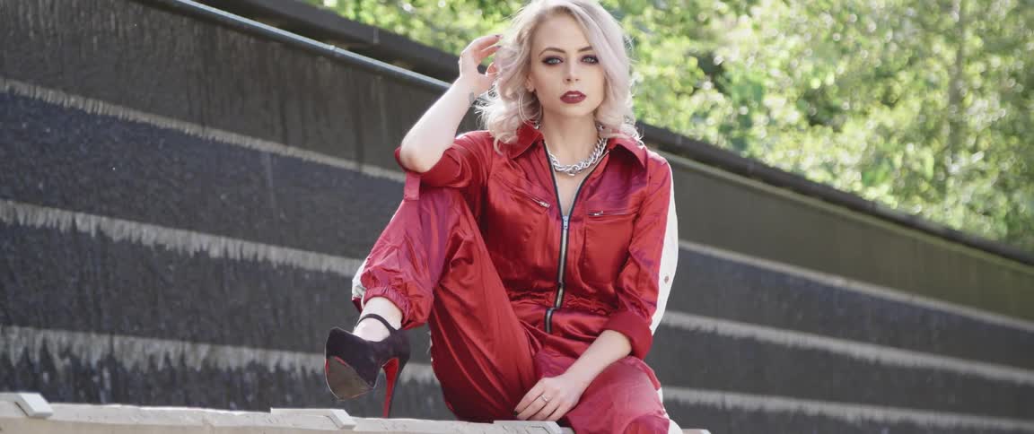 A woman wearing a red jumpsuit poses sitting with her leg up and high heels on display