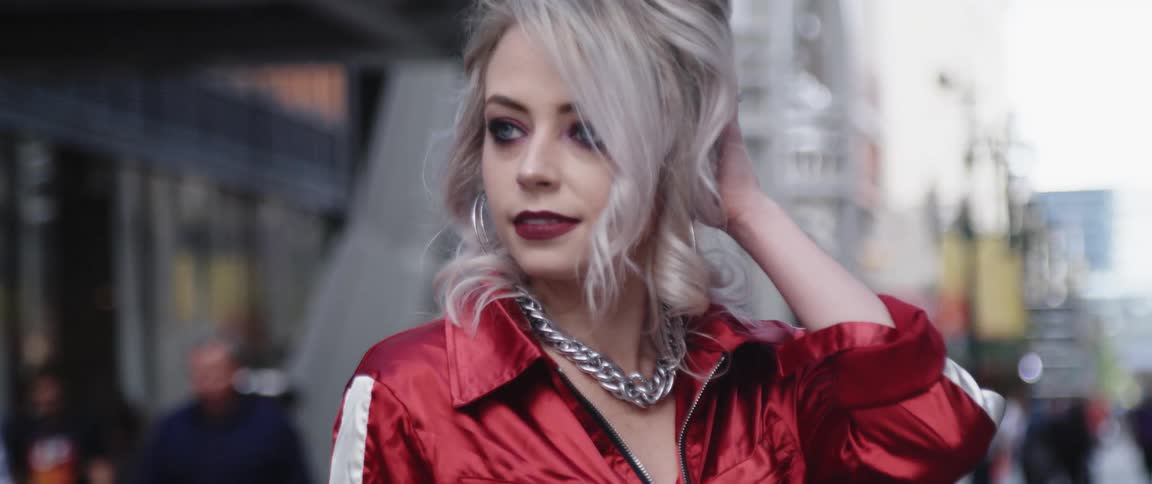 Female model poses in red silky top and large chain necklace as pedestrians pass on street in the background