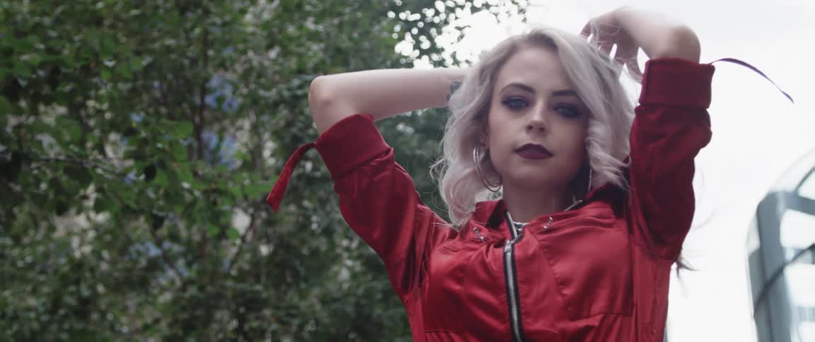 Woman with makeup gray hair hoop earrings and red jacket looks down at camera in city street