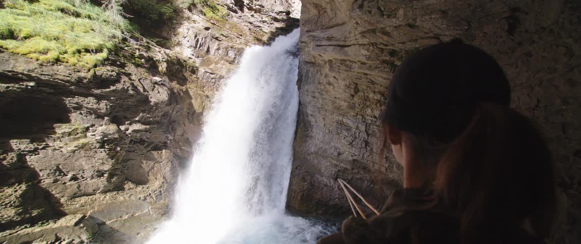 Woman stands in cavern watching river flow down waterfall across from grass topped incline