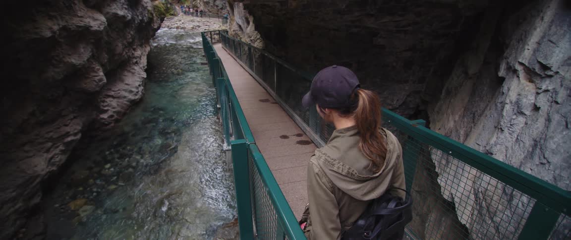 Woman walks down inclined walkway between shoulder hide barriers looking down toward river sitting at bottom of ravine