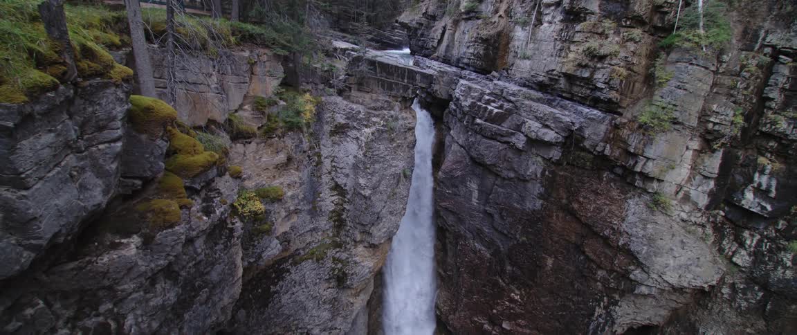 Woman wearing baseball cap stands on observation platform looking across to waterfall falling down side of cliff topped by conifer trees