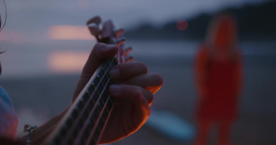 Woman plays ukulele next to bonfire on the beach at dusk
