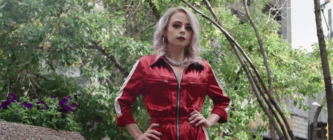 A young blonde haired woman wearing a red jumpsuit has her hands on her hips as she walks with trees and a building behind her