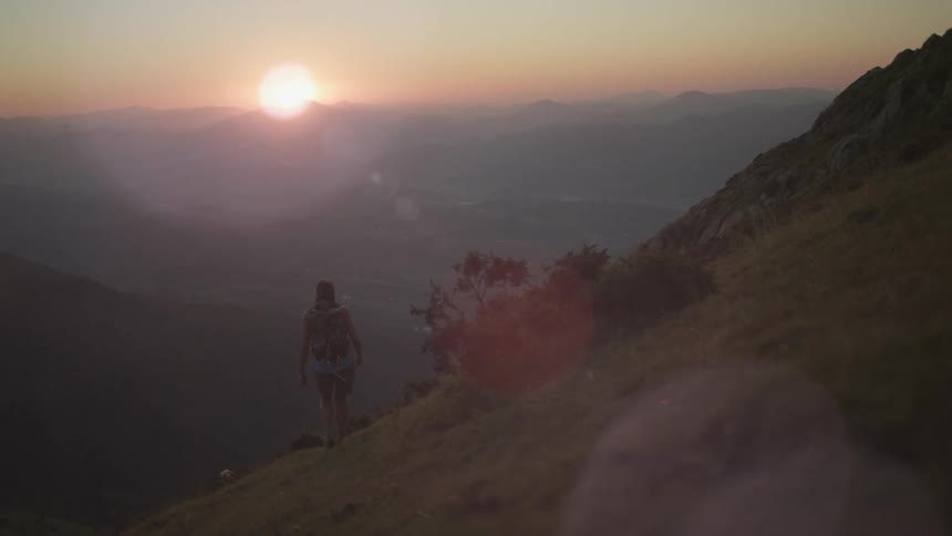 A hiker wearing a backpack walks down the side of a mountain as the sun shines brightly over mountain tops on the horizon