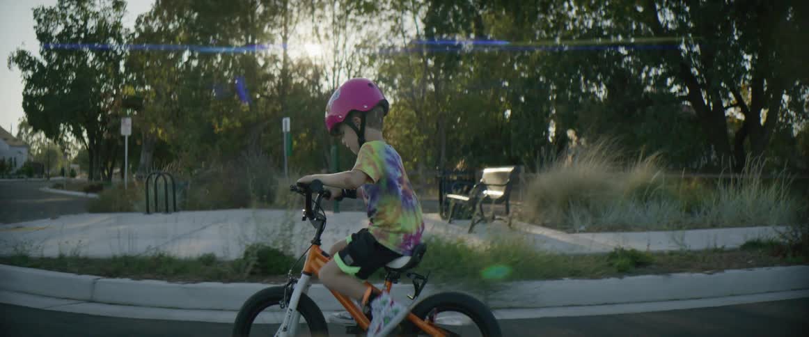 Child wearing helmet pedals bicycle with training wheels along neighborhood street passing bench on corner