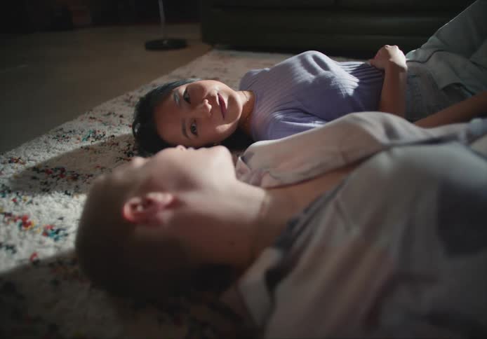 A pair of women talk with one another while lying on a rug indoors during the day