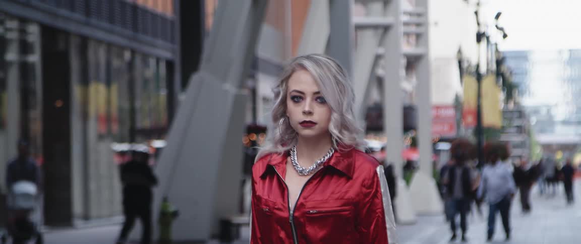 Model wearing large silver necklace and red top moves forward as pedestrians walking along city street in background