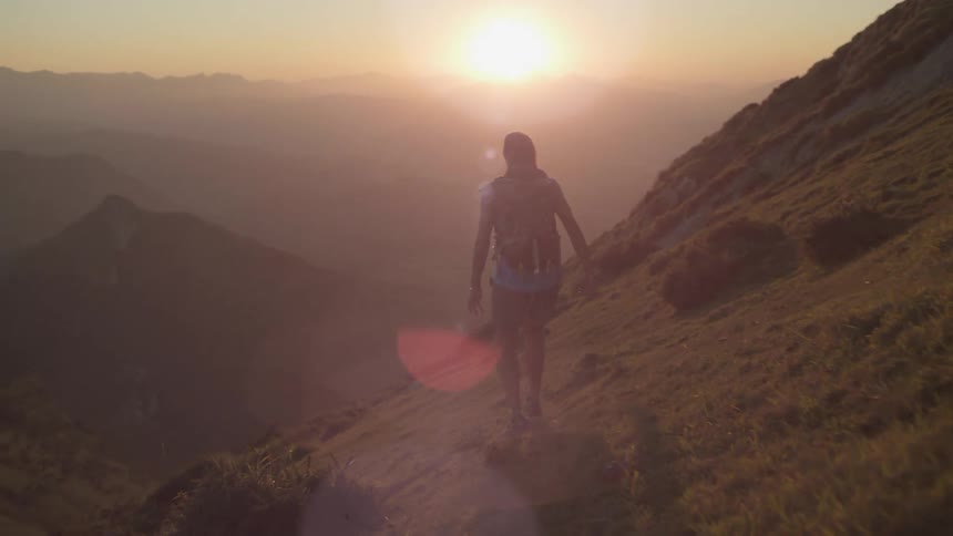 Man with backpack shorts and sleeveless shirt walks on mountains slope at sunrise