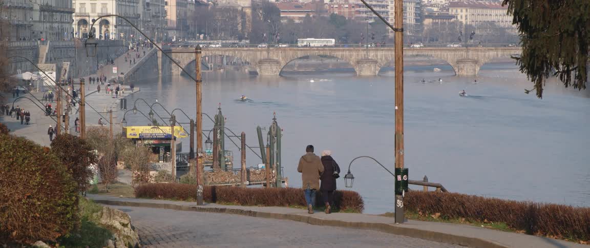 A couple walk arm in arm on a waterfront sidewalk heading towards the city and a traffic covered bridge