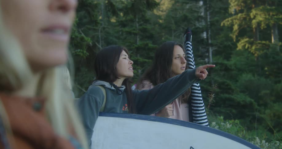 Woman carrying a surfboard standing in the forest with friends points at the horizon