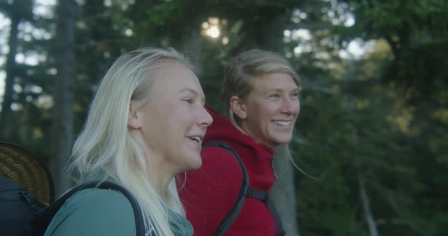 Two women with backpacks on stand in a forest and talk and laugh as one woman points in the distance