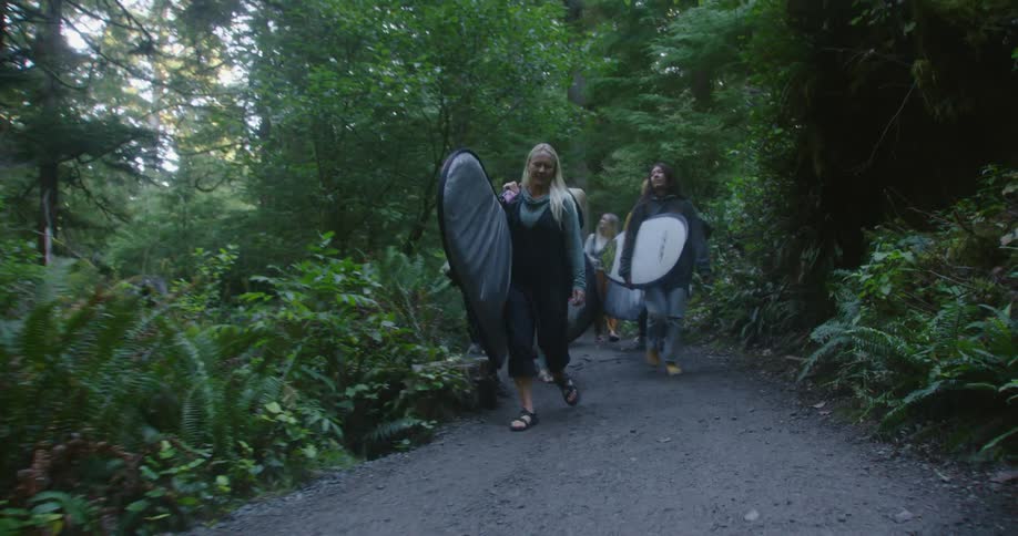 Female friends carry surfboards in bags down path through woods as they smile and talk