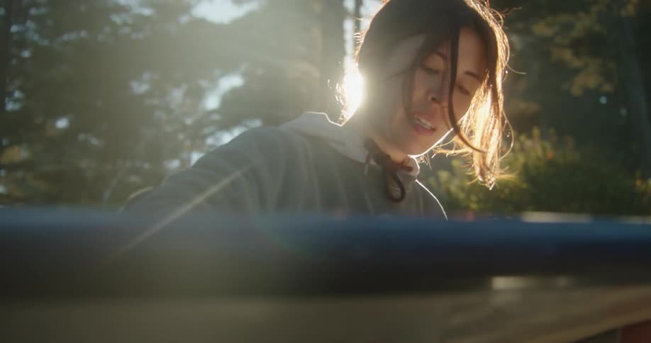 Young woman sitting on the forest ground waxing a surfboard at morning