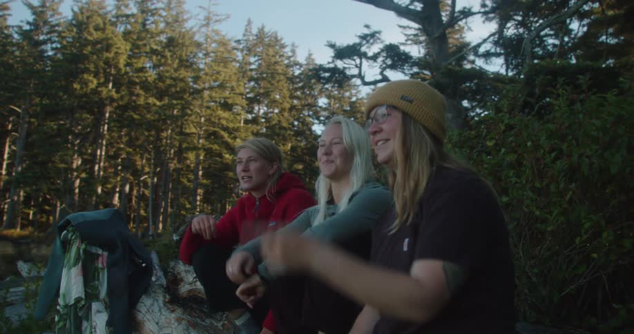 Female friends sit on log pointing and gesturing as they talk near conifer forest