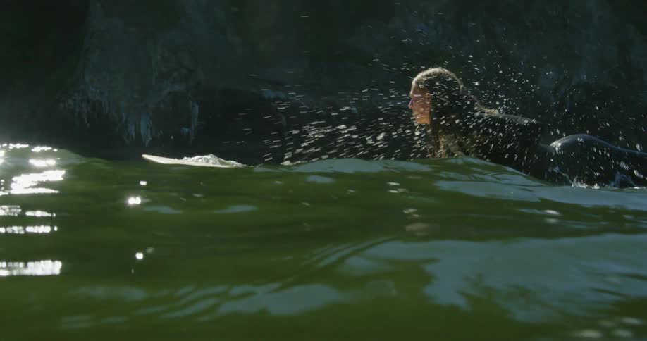 A girl wearing a wet suit is on her stomach paddling her surfboard with her hands