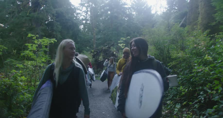 A group of women carry surfboards while walking through a woodland during the day