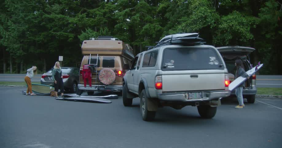 Three women with surfboards in a parking lot wave as a truck pulls in