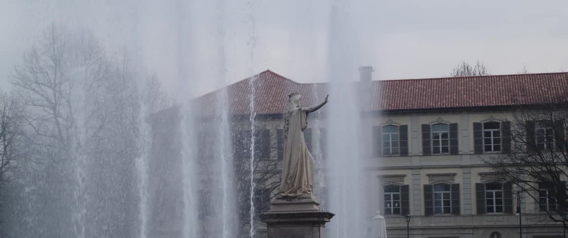 Fountain spouts water high into the air around centrally placed statue with red roofed building in the background