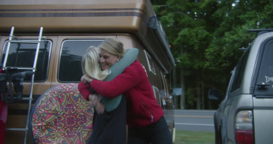 Woman gets out of vehicle with boat on the roof to greet two other women one from a van with a camper storage on the roof