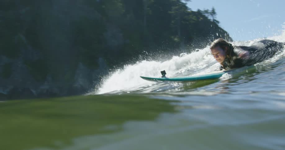 A woman lying atop a surfboard begins to stand as an ocean wave catches up with her