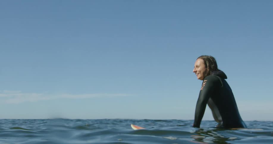 A young woman wearing a wet suit is sitting on a surfboard looking around at the water