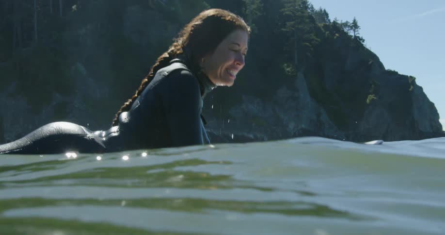 Female surfers paddle out to sea on surfboards bobbing in the water with forest topped cliff in background