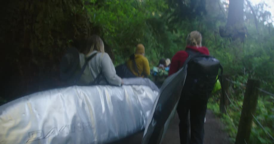 Women carrying surfboards in bags move along path through woods