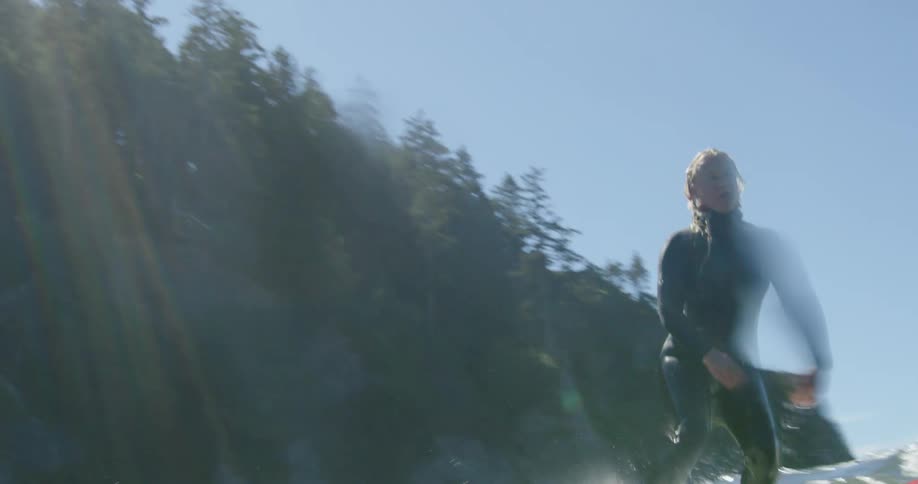 Young blonde woman wearing black wet suit rides a surfboard on the waves near the mountains