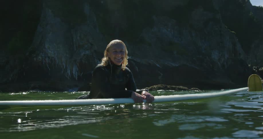 A woman smiles as she poses while leaning onto her surfboard in the ocean