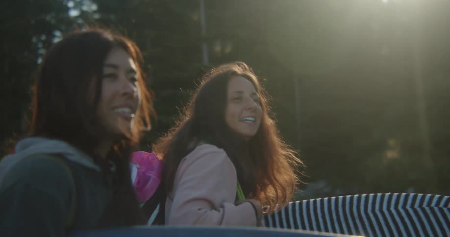 Two female surfers carrying surfboard walk by area of land covered with trees