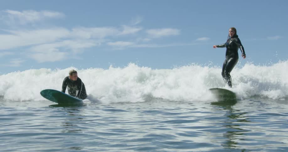 A woman surfs awkwardly towards the beach alongside another woman