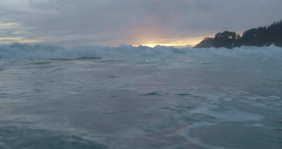 Rough water and waves roll in with the sun on the horizon in a cloudy sky