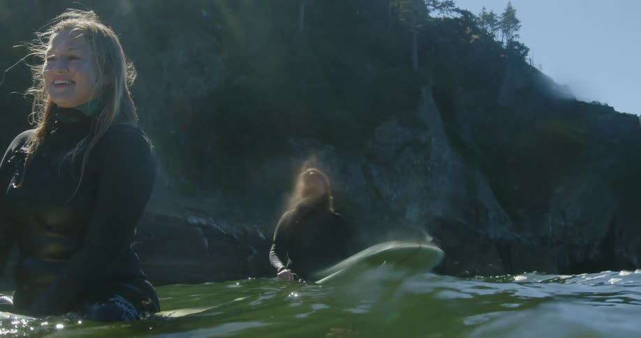 A group of women sit atop their surfboards on the ocean beneath a cliff during the day