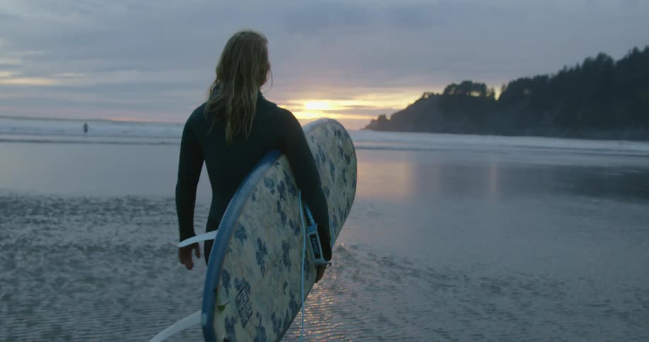 Woman with blond wavy hair and wetsuit carrying surfboard walks on the beach at sunset