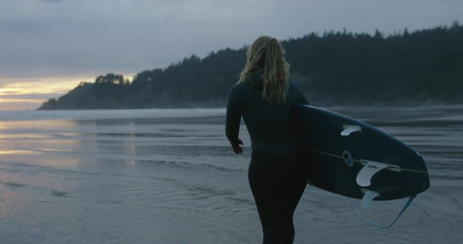 Female surfer carries surfboard across beach as sun sits just above the horizon on a misty day