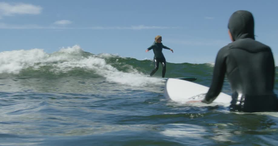 A surfer stands on her surfboard riding a wave as another surfer is in the water nearby