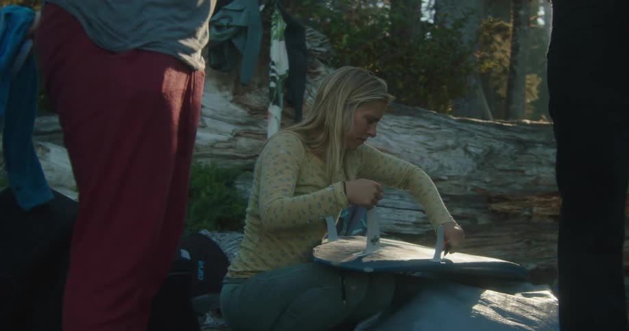 A woman knees on the ground and tightens fins on a surfboard as she talks with two other people