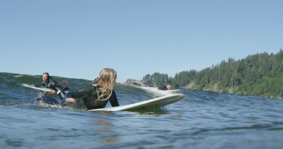 Blonde young woman wearing a black wet suit riding a surfboard over the ocean waves near the mountains