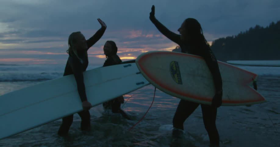 A group of woman high five standing in shallow ocean water and holding surfboards